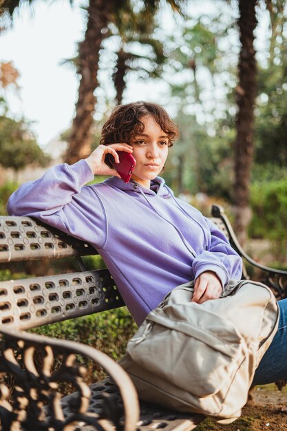 Jovencita sentada en un banco de un parque hablando con un amigo por teléfono.