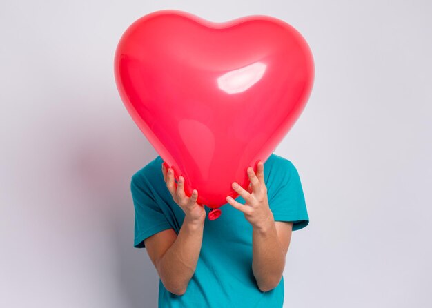 Jovencito escondido detrás de un globo en forma de corazón rojo Niño sosteniendo el símbolo del amor en el estudio