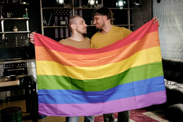 Foto jóvenes afectuosos hombres homosexuales sosteniendo la bandera del arco iris y mirando el uno al otro cuando están de pie en el apartamento