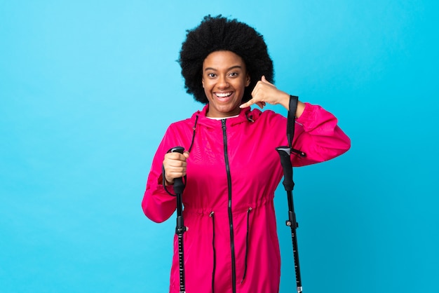 Jóvenes africanos americanos con mochila y bastones de trekking aislados en azul haciendo gesto de teléfono. Llámame señal de vuelta