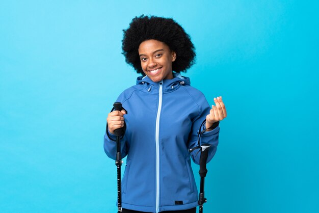 Jóvenes africanos americanos con mochila y bastones de trekking aislados en la pared azul haciendo dinero gesto