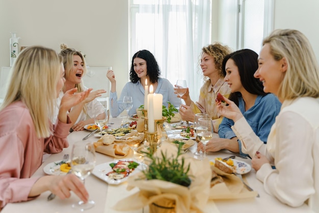 Jóvenes amigas felices bebiendo vino y riéndose de la mesa festiva servida con comida fresca y decorada con velas