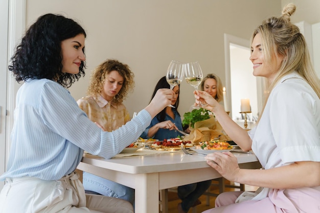 Jóvenes amigas felices tintineando vasos y sonriendo en la mesa festiva servida con comida y vino blanco decorado con velas