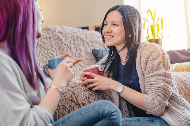Jóvenes amigas sonrientes con tazas de té hablando mientras se sientan en el sofá en casa