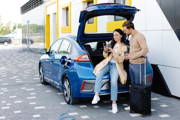 Jóvenes amigos cargando la batería del automóvil desde la estación pública y parados cerca del estacionamiento para un automóvil eléctrico Hombre y mujer hablando y bebiendo café en la ciudad