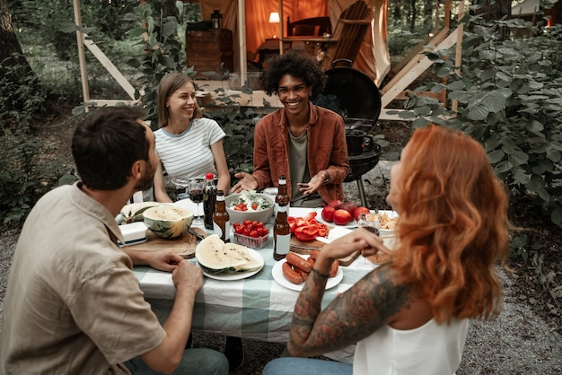 Jóvenes amigos cenando en glamping clincking gafas después del atardecer. Feliz pandilla milenaria acampando en un picnic al aire libre bajo las luces de la bombilla. Pasar tiempo con amigos al aire libre, fiesta de barbacoa.