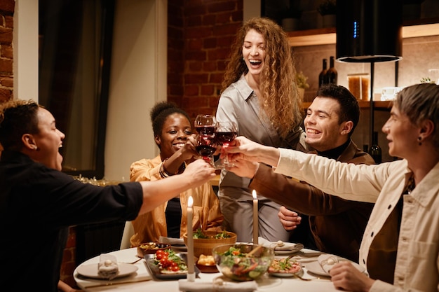 Jóvenes amigos extasiados tintineando con vino tinto en copas de vino sobre la mesa servida con comida casera