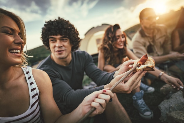 Jóvenes amigos felices disfrutan de un agradable día soleado en la naturaleza. Están asando salchichas, comiendo, riendo y hablando felices de estar juntos.