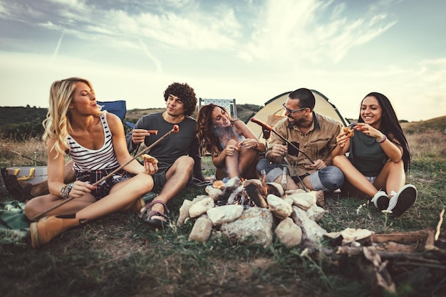 Jóvenes amigos felices disfrutan de un buen día en la naturaleza. Están asando salchichas, comiendo, riendo y hablando felices de estar juntos.