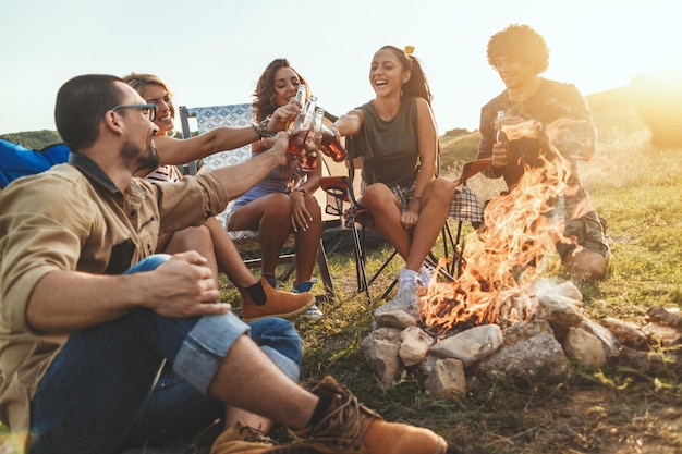 Jóvenes amigos felices disfrutan de un día soleado en la montaña. Están riendo y brindando con botellas de cerveza junto a la fogata felices de estar juntos.