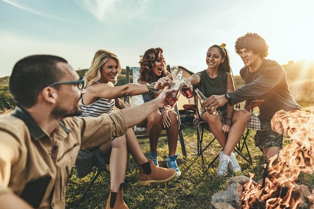 Jóvenes amigos felices disfrutan de un día soleado en la montaña. Están riendo y brindando con botellas de cerveza junto a la fogata felices de estar juntos.