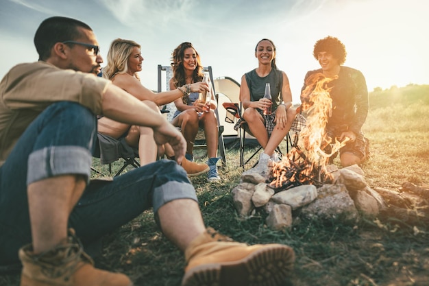 Jóvenes amigos felices disfrutan de un día soleado en la montaña. Se ríen y brindan con botellas de cerveza junto a la hoguera cerca de la tienda.