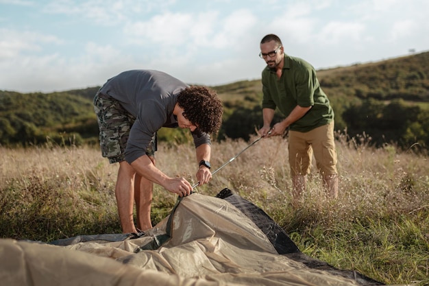 Los jóvenes amigos felices se están preparando para acampar. Están instalando una tienda de campaña en un lugar adecuado en un prado.