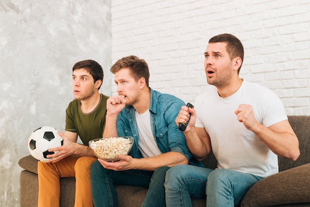 Jóvenes amigos viendo partido de fútbol en televisión con expresiones serias.