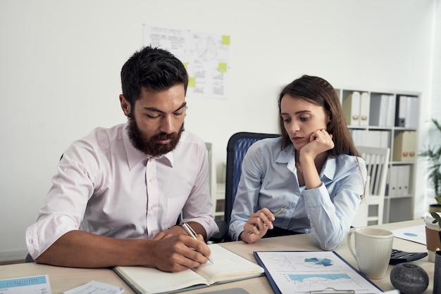 Jóvenes colegas caucásicos concentrados sentados en la mesa y tomando notas en el diario mientras discuten ideas de negocios