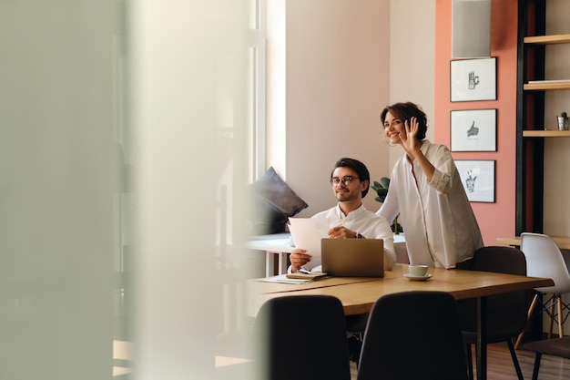 Jóvenes colegas de negocios sonrientes con papeles saludando felizmente a alguien en el trabajo en una oficina moderna