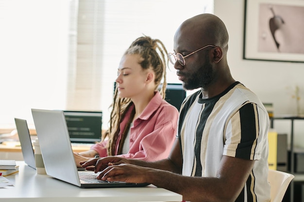 Jóvenes colegas sentados en la mesa y escribiendo códigos en computadoras durante su trabajo en la oficina