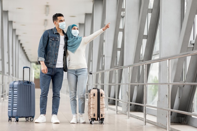 Jóvenes cónyuges musulmanes con mascarillas médicas de pie con equipaje en el aeropuerto