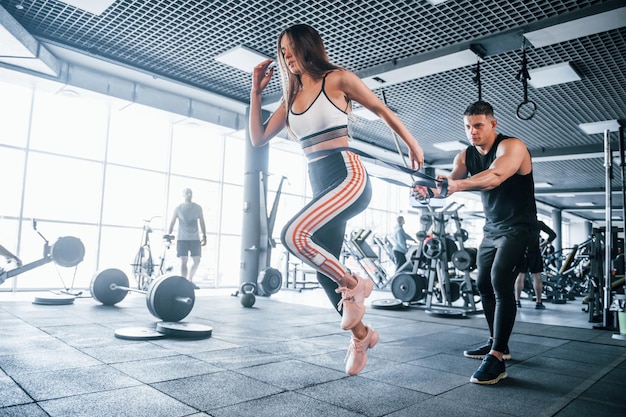 Jóvenes deportistas haciendo ejercicios juntos en el gimnasio Mujer corriendo cuando el hombre la sostiene con cinta elástica