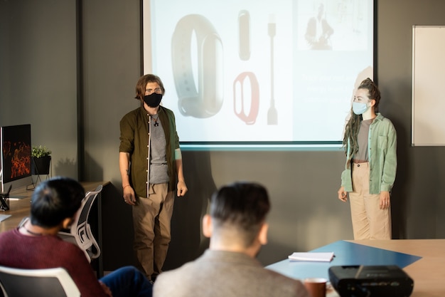 Jóvenes diseñadores confiados en ropa casual y máscaras protectoras de pie junto a la pizarra durante la presentación de nuevos modelos de smartwatch