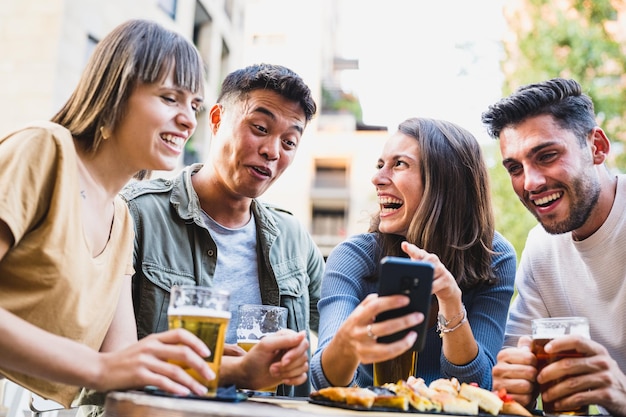 Jóvenes divirtiéndose en la cervecería una mujer riendo bromeando con amigos mirando un teléfono inteligente joven asiático haciendo una cara graciosa estilo de vida de los millennials felicidad y concepto despreocupado