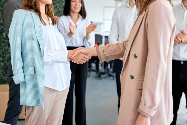 Jóvenes dos señoras de negocios recortadas en ropa formal estrecharme la mano contrato de acabado en oficina moderna y luminosa en presencia de colegas aplaudiendo. Apretón de manos y marketing. Copie el espacio. Vista lateral