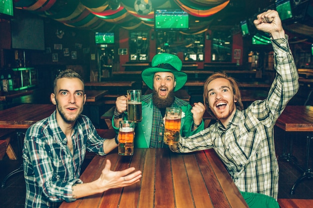 Los jóvenes emocionados se sientan a la mesa en el pub y miran hacia adelante. Ellos animan Los hombres sostienen jarras de cerveza en las manos. Hombre joven en medio llevar traje verde de San Patricio.