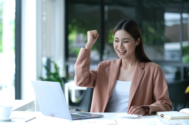 Las jóvenes empresarias asiáticas muestran una expresión alegre de éxito en el trabajo sonriendo felizmente con una computadora portátil en una oficina moderna