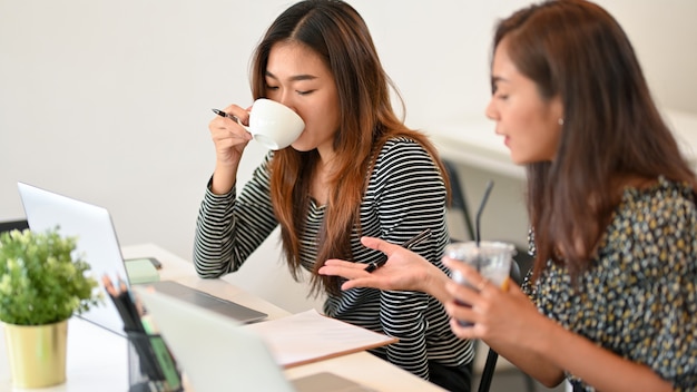 Jóvenes empresarias asiáticas que trabajan con una computadora portátil y beben café en el espacio de coworking de café