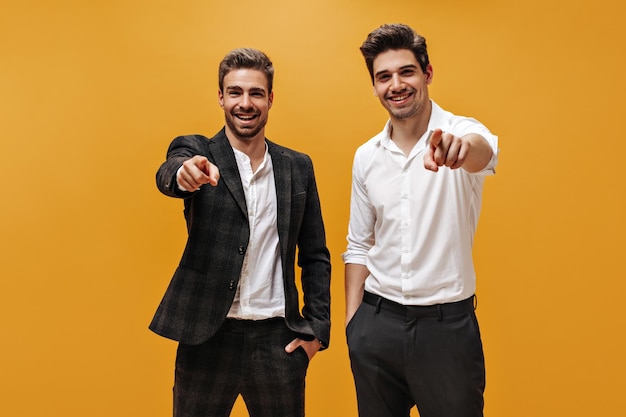 Jóvenes empresarios brunet de buen humor con camisas blancas y pantalones negros apuntan a la sonrisa de los abdominales de la cámara en un fondo naranja aislado