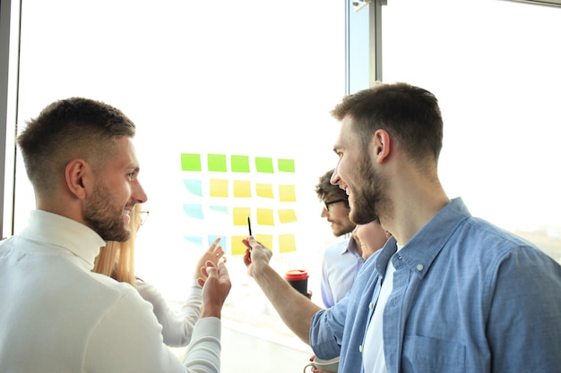 Foto jóvenes empresarios creativos de inicio en reunión en la oficina moderna haciendo proyectos de planes con pegatinas de poste sobre vidrio.