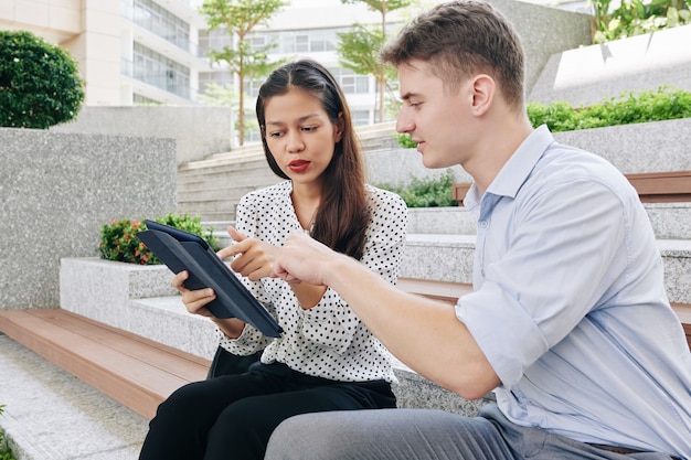 Jóvenes empresarios multiétnicos discutiendo el proyecto en la tableta cuando descansan sobre los pasos al aire libre durante las vacaciones