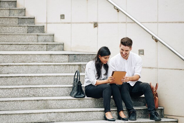 Jóvenes empresarios multiétnicos sentados en los pasos y leyendo el artículo en la tableta