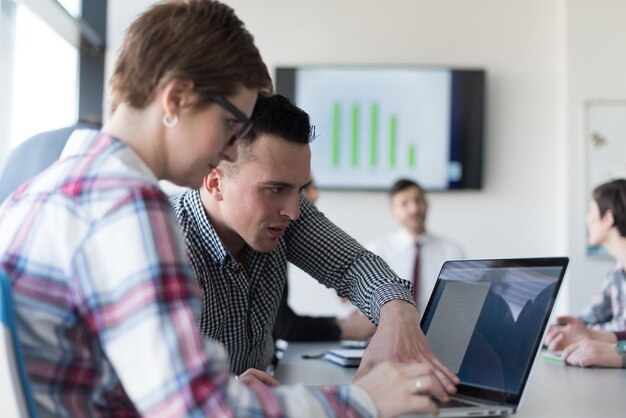 jóvenes empresarios de nueva creación, pareja que trabaja en una computadora portátil, grupo de empresarios reunidos en segundo plano en el interior de la oficina