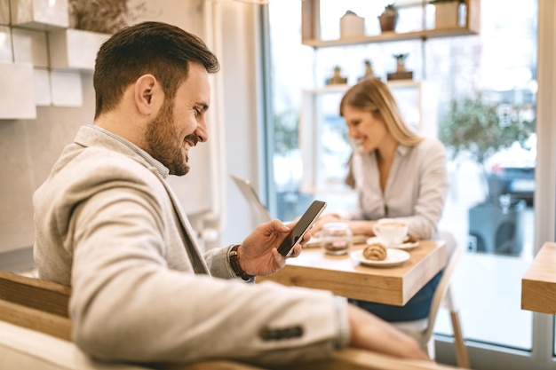 Jóvenes empresarios sonrientes en un descanso en un café. Hombre que usa un teléfono inteligente y lee un mensaje de Internet. Mujer trabajando en tableta.