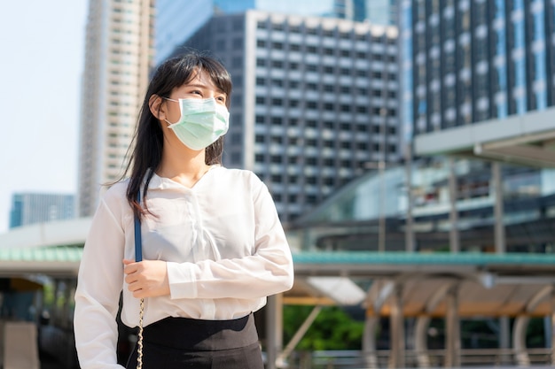 Jóvenes estresan a una empresaria asiática con camisa blanca que va a trabajar en una ciudad contaminada que usa una máscara de protección para evitar el polvo y COVID-19 con un edificio de oficinas comerciales en Bangkok, Tailandia.