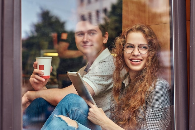 Jóvenes estudiantes felices bebiendo café y usando una tableta digital sentados en un umbral de ventana en un campus universitario durante un descanso.