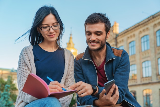 Jóvenes estudiantes sentados al aire libre mirando el teléfono móvil mientras sonríen y toman notas