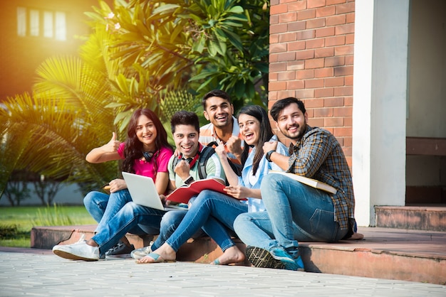 Jóvenes estudiantes universitarios indios asiáticos leyendo libros, estudiando en la computadora portátil, preparándose para el examen o trabajando en un proyecto grupal mientras están sentados en el césped, la escalera o los escalones del campus universitario