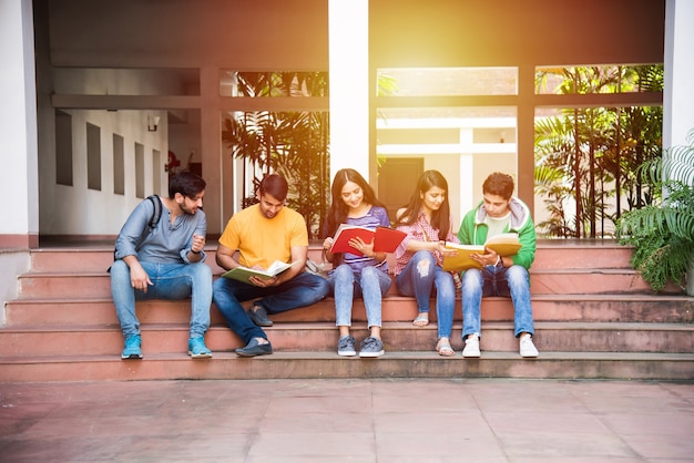 Jóvenes estudiantes universitarios indios asiáticos leyendo libros, estudiando en la computadora portátil, preparándose para el examen o trabajando en un proyecto grupal mientras están sentados en el césped, la escalera o los escalones del campus universitario