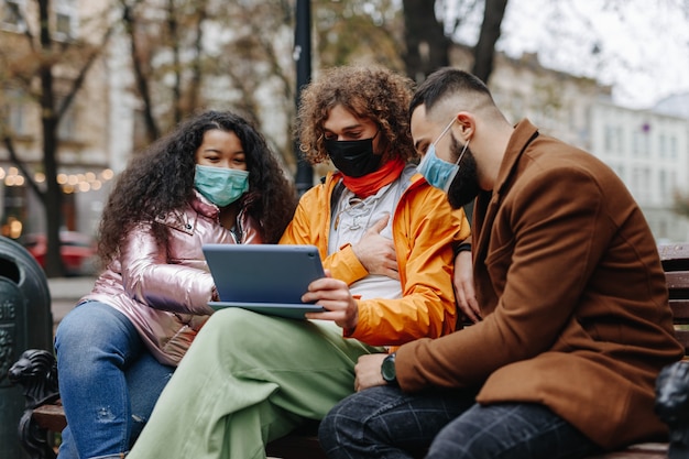 Jóvenes felices con tableta digital mientras pasan tiempo libre al aire libre. Personas multiculturales con máscaras médicas sentados en un banco de madera al aire libre.