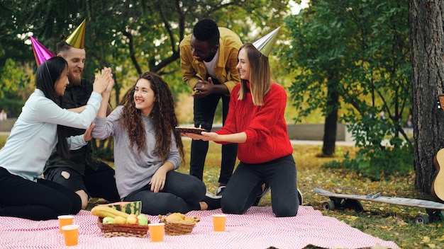 Los jóvenes felicitan a la niña por su cumpleaños trayendo pastel riéndose y regocijándose durante una fiesta al aire libre en el parque