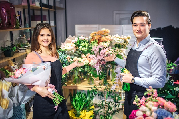 Jóvenes floristas masculinos y femeninos de pie y mirar. Ella sostiene un ramo de flores. Tiene agua pulverizada y punto en flores. Se ven bonitas y alegres.