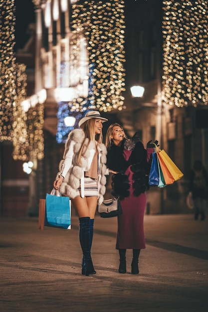 Jóvenes hermosas dos hermanas alegres, con coloridas bolsas de compras, divirtiéndose y caminando por la calle de la ciudad de noche en Navidad.