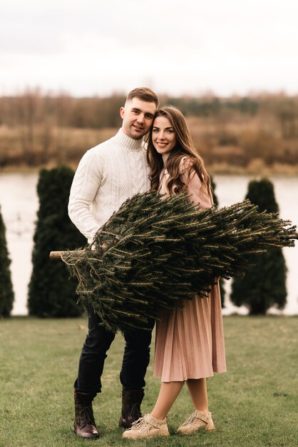 Jóvenes hermosos amantes felices hombre y mujer, historia de amor en invierno con un árbol de Navidad vivo en sus manos en el fondo de la orilla del río