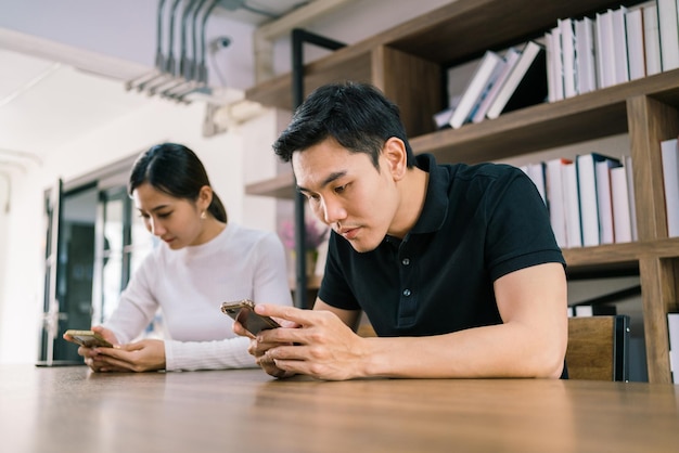 Jóvenes hombres y mujeres asiáticos trabajando juntos en la mesa de madera con una laptop, mirando sus teléfonos móviles con una estantería en el fondo.