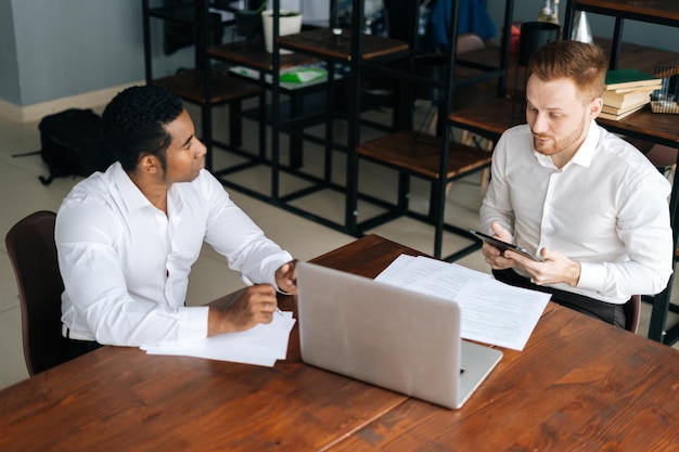 Jóvenes hombres de negocios europeos afroamericanos y caucásicos que usan ropa formal sentados juntos en el escritorio de la oficina con documentos en papel de computadora portátil tableta digital y discutiendo un nuevo proyecto o idea de negocio