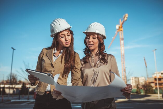 Foto jóvenes ingenieras exitosas con cascos blancos y planos en sus manos están analizando un nuevo proyecto en el sitio de construcción.