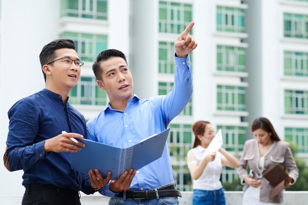 Jóvenes inversores vietnamitas comprobando documentos y mirando edificios alrededor cuando están parados en la azotea del edificio de oficinas