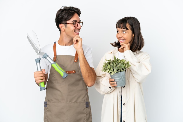 Jóvenes jardineros de raza mixta sosteniendo una planta y tijeras de podar aisladas de fondo blanco mirándose el uno al otro